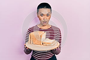 Beautiful hispanic woman with short hair holding sandwich bread in shock face, looking skeptical and sarcastic, surprised with