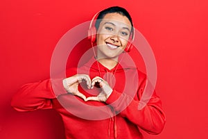 Beautiful hispanic woman with short hair listening to music using headphones smiling in love doing heart symbol shape with hands