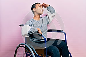 Beautiful hispanic woman with short hair sitting on wheelchair very happy and smiling looking far away with hand over head