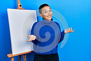 Beautiful hispanic woman with short hair standing by painter easel stand clueless and confused expression with arms and hands