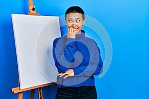 Beautiful hispanic woman with short hair standing by painter easel stand looking stressed and nervous with hands on mouth biting