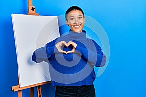 Beautiful hispanic woman with short hair standing by painter easel stand smiling in love showing heart symbol and shape with hands