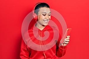 Beautiful hispanic woman with short hair using smartphone wearing headphones looking positive and happy standing and smiling with