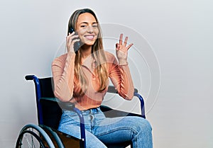 Beautiful hispanic woman sitting on wheelchair talking on the phone doing ok sign with fingers, smiling friendly gesturing