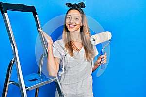 Beautiful hispanic woman by stairs holding roller painter celebrating victory with happy smile and winner expression with raised