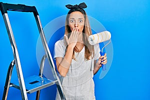 Beautiful hispanic woman by stairs holding roller painter covering mouth with hand, shocked and afraid for mistake