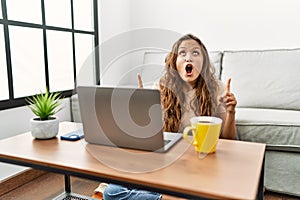 Beautiful hispanic woman using computer laptop at home amazed and surprised looking up and pointing with fingers and raised arms