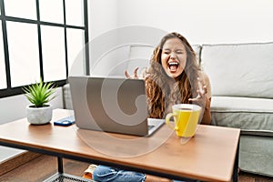 Beautiful hispanic woman using computer laptop at home celebrating mad and crazy for success with arms raised and closed eyes