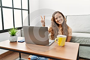 Beautiful hispanic woman using computer laptop at home smiling with happy face winking at the camera doing victory sign