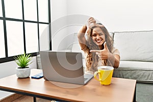 Beautiful hispanic woman using computer laptop at home smiling making frame with hands and fingers with happy face
