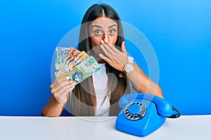 Beautiful hispanic woman with vintage telephone holding australian dollars covering mouth with hand, shocked and afraid for