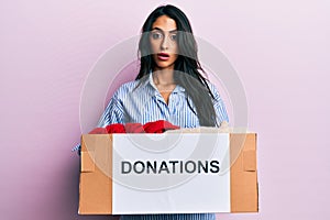 Beautiful hispanic woman volunteer holding donations box in shock face, looking skeptical and sarcastic, surprised with open mouth