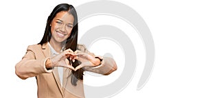 Beautiful hispanic woman wearing business jacket smiling in love doing heart symbol shape with hands