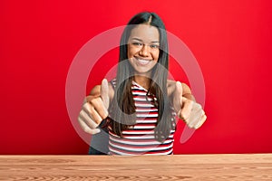 Beautiful hispanic woman wearing casual clothes sitting on the table approving doing positive gesture with hand, thumbs up smiling