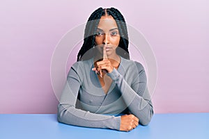 Beautiful hispanic woman wearing casual clothes sitting on the table asking to be quiet with finger on lips