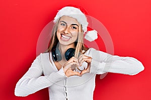 Beautiful hispanic woman wearing christmas hat and headphones smiling in love doing heart symbol shape with hands