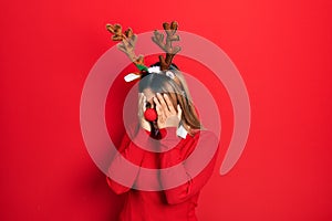 Beautiful hispanic woman wearing deer christmas hat and red nose with sad expression covering face with hands while crying