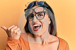 Beautiful hispanic woman wearing many glass over face pointing thumb up to the side smiling happy with open mouth