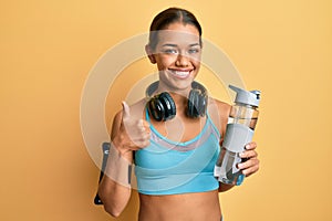 Beautiful hispanic woman wearing sportswear drinking bottle of water smiling happy and positive, thumb up doing excellent and