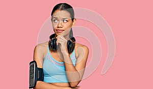 Beautiful hispanic woman wearing sportswear and headphones serious face thinking about question with hand on chin, thoughtful