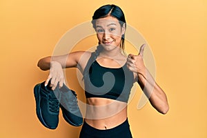 Beautiful hispanic woman wearing sportswear showing sporty shoes smiling happy and positive, thumb up doing excellent and approval