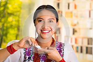 Beautiful hispanic woman wearing white blouse with colorful embroidery, applying cream onto face using finger during