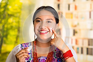 Beautiful hispanic woman wearing white blouse with colorful embroidery, applying cream onto face using finger during