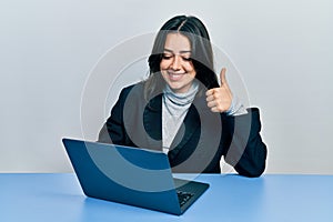 Beautiful hispanic woman working at the office with laptop smiling happy and positive, thumb up doing excellent and approval sign