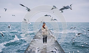 Beautiful horizontal image of young woman feeds seagulls on the sea. Pretty female wearing coat, scarf and watching flying