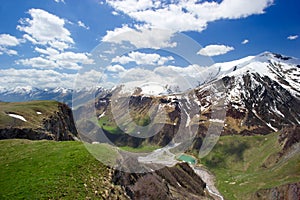 Beautiful huge mountains peaks in snow, blue sky, white clouds, blue lake at the foot of the mountain, green hills, green valley