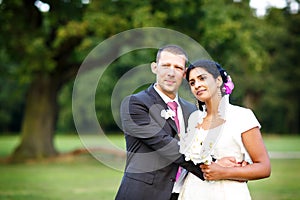 Beautiful indian bride and caucasian groom, in summer park. Happy young woman holding flowers. Young man smiling, Couple