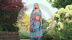 Beautiful Indian fantasy woman posing in blooming spring nature park. Oriental girl in national pink blue sari dress