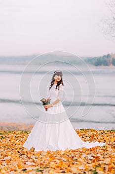 Beautiful innocent thoughtful bride in gorgeous white dress stands on fallen leaves at riverside