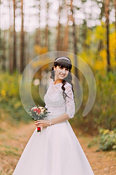 Beautiful innocent young brunette bride in classic white dress stands on forest trail