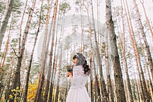 Beautiful innocent young brunette bride in gorgeous white dress with bouquet at pine wood forest