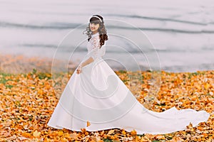 Beautiful innocent young brunette bride in gorgeous white dress stands on fallen leaves at riverside