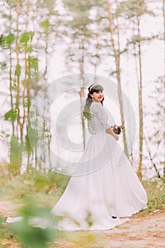 Beautiful innocent young brunette bride in gorgeous white dress stands on forest trail at sunny day