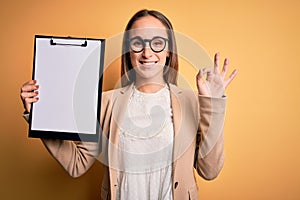 Beautiful inspector woman wearing glasses holding checklist clipboard over yellow background doing ok sign with fingers, excellent