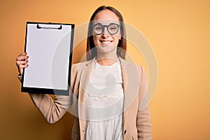 Beautiful inspector woman wearing glasses holding checklist clipboard over yellow background with a happy face standing and