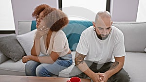 Beautiful interracial couple sitting in silence on living room sofa, stress and anger visible in expression, undercurrent of