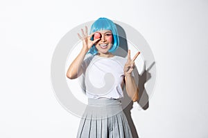 Beautiful japanese girl in blue wig and schoolgirl costume holding macaron, showing peace gesture and smiling, standing