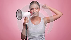 A beautiful joyful girl in a striped T-shirt shouts into the loudspeaker on isolated pink background