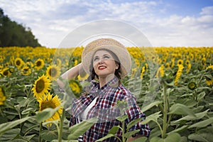 Beautiful joyful and smiling middle-aged farmer woman in a straw hat and shirt stands in a harvest field of sunflowers on a sunny