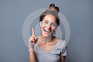Beautiful joyful woman with a natural open smile pointing with index finger upwards over gray background