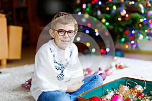 Beautiful kid boy with eye glasses and colorful vintage xmas toys and balls in old suitcase. Child decorating Christmas