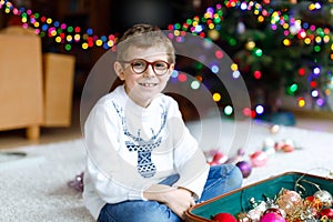 Beautiful kid boy with eye glasses and colorful vintage xmas toys and balls in old suitcase. Child decorating Christmas