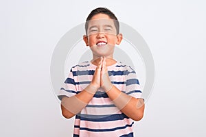 Beautiful kid boy wearing casual striped t-shirt standing over isolated white background begging and praying with hands together