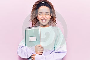 Beautiful kid girl with curly hair holding notebook looking positive and happy standing and smiling with a confident smile showing
