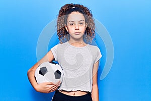 Beautiful kid girl with curly hair holding soccer ball thinking attitude and sober expression looking self confident