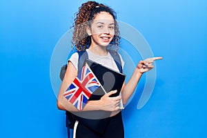 Beautiful kid girl with curly hair wearing student backpack holding binder and uk flag smiling happy pointing with hand and finger